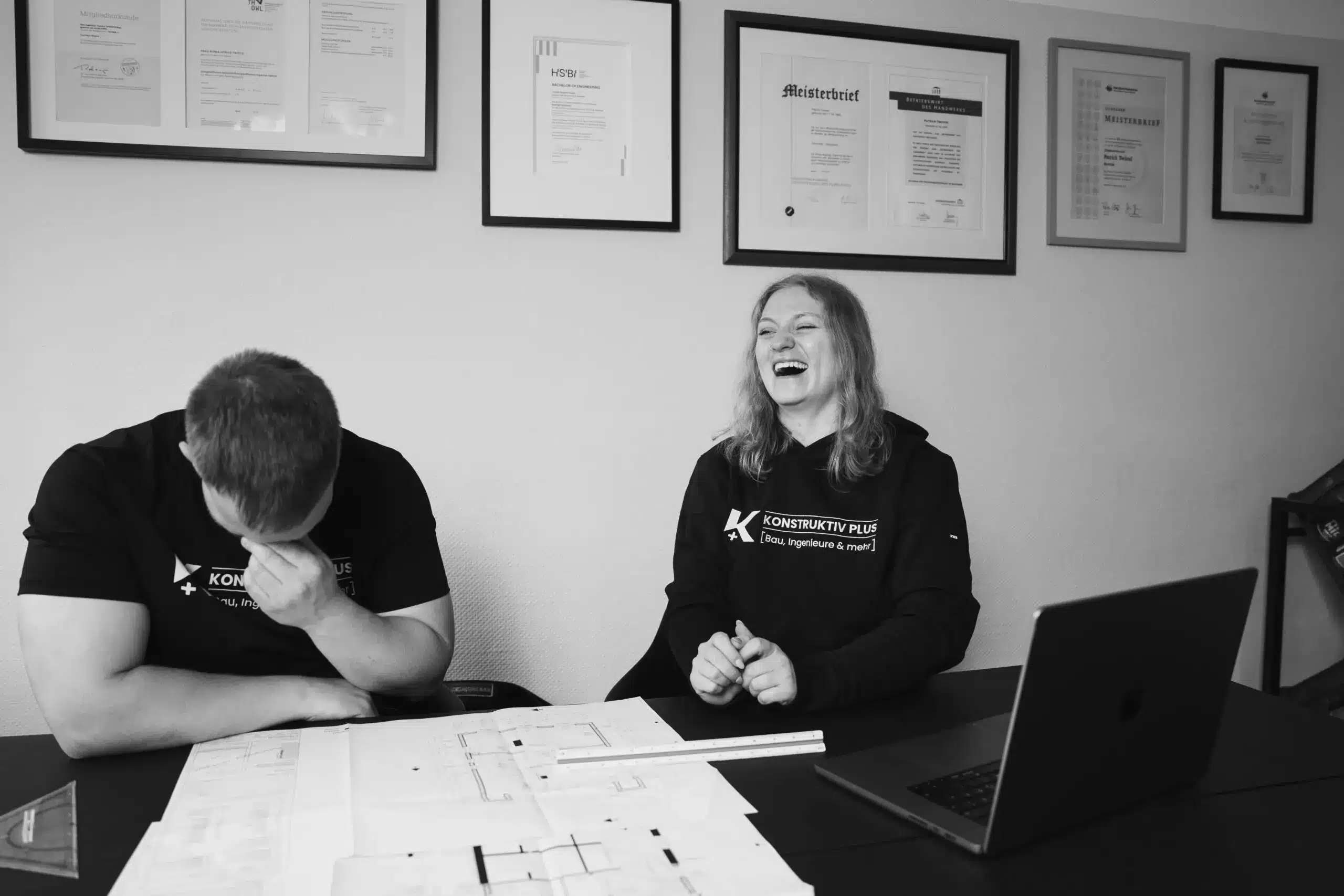 Yannick und Ronja Twistel von Konstruktiv Plus im schwarzen Firmen-Shirt sitzt einem Kollegen gegenüber und spricht mit ihm – auf dem Tisch liegen Baupläne und ein Laptop.