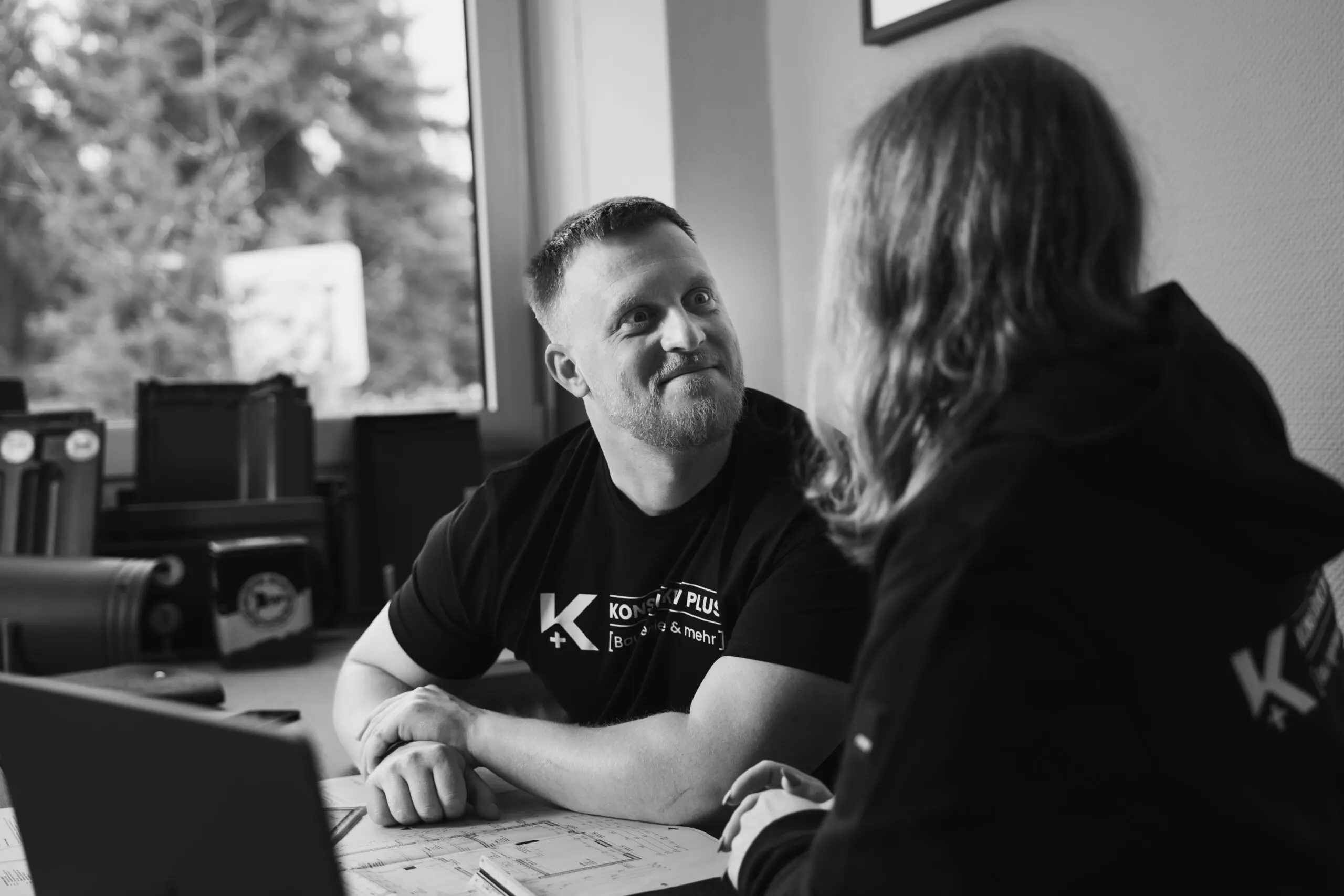 Yannick und Ronja Twistel von Konstruktiv Plus im schwarzen Firmen-Shirt sitzt einem Kollegen gegenüber und spricht mit ihm – auf dem Tisch liegen Baupläne und ein Laptop.