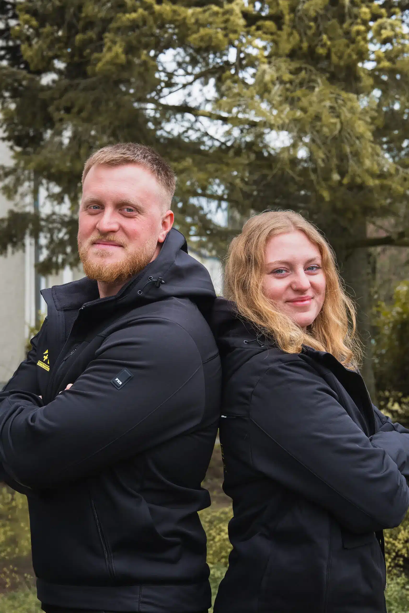 Yannick und Ronja Twistel von Konstruktiv Plus stehen Rücken an Rücken im Freien und blicken selbstbewusst in die Kamera.