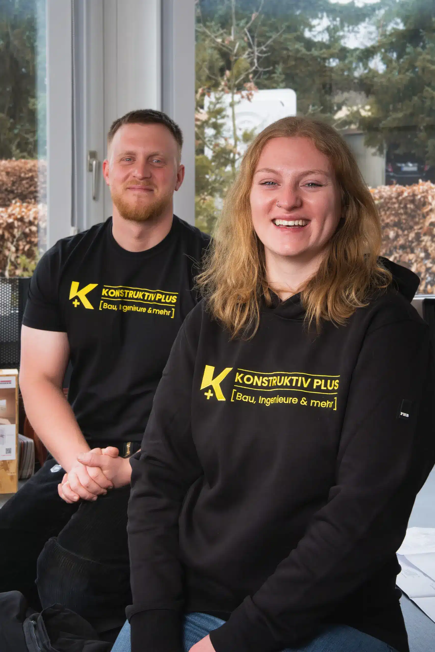 Yannick und Ronja Twistel von Konstruktiv Plus sitzen im Büro und lächeln in die Kamera, tragen schwarze Kleidung mit Firmenlogo.